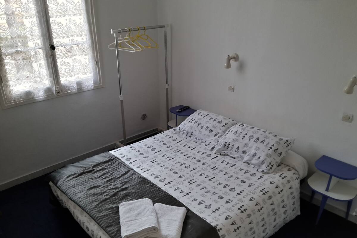 Un dormitorio con cama, mesita de noche, perchero y ventana con cortinas de encaje.