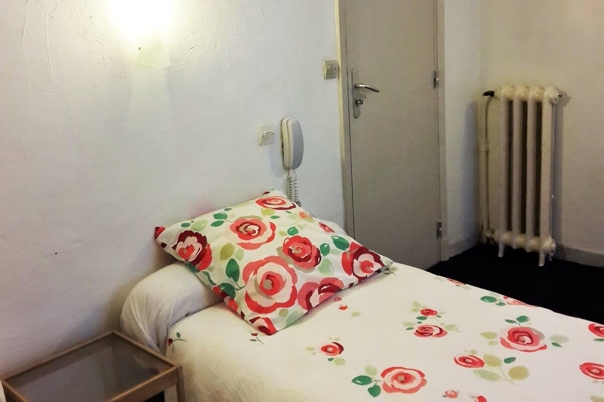 Una habitación pequeña con cama individual, colcha estampada, mesita de noche, teléfono en la pared y radiador.