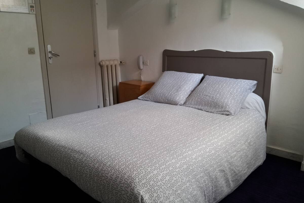 Una cama bien hecha con ropa de cama estampada, cabecera de madera, mesita de noche con lámpara y radiador.