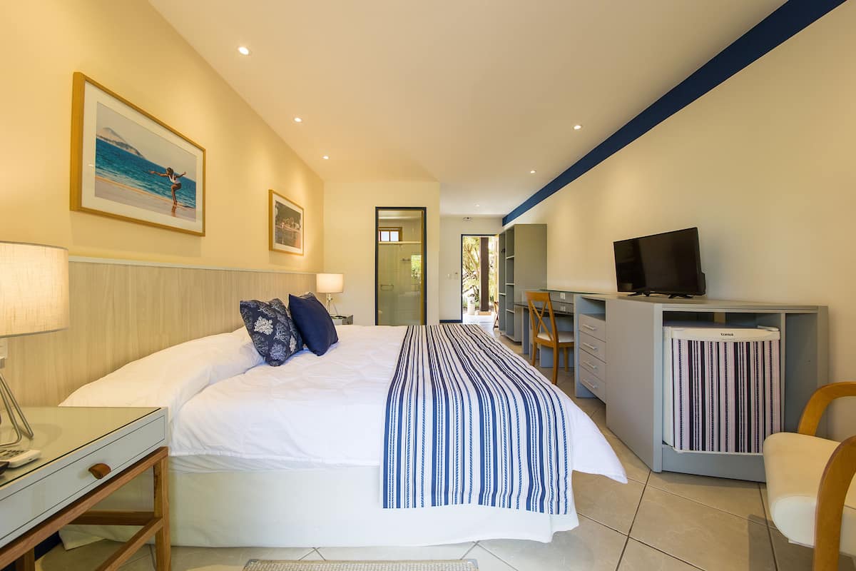 A hotel room with a bed, bedside table, lamp, TV, and a desk with a chair.