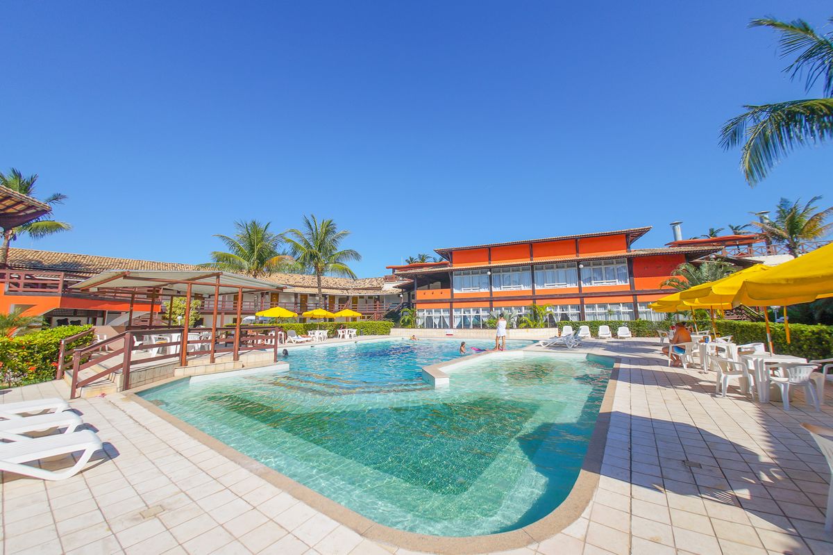 A swimming pool with clear water, surrounded by lounge chairs and umbrellas, and a building with orange and red exterior walls.