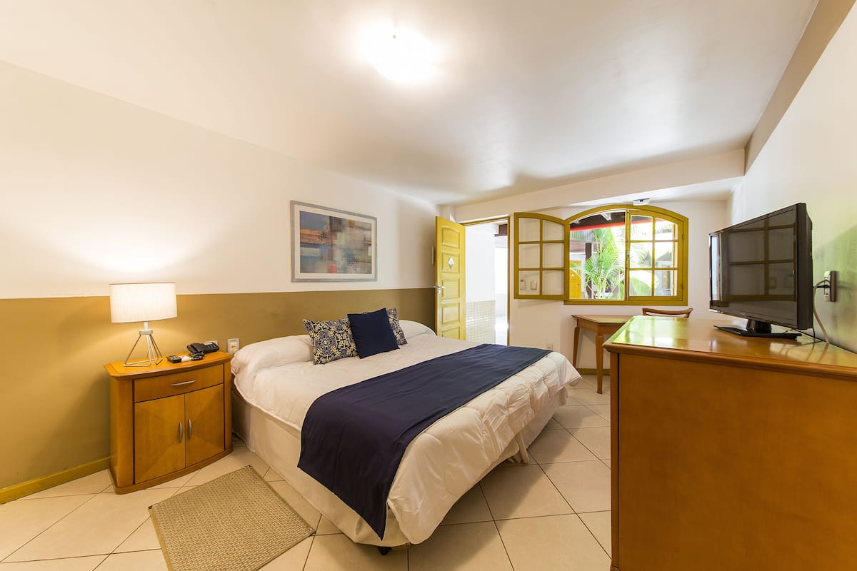 A hotel room with a bed, bedside table, lamp, TV, and a window with a view of greenery.
