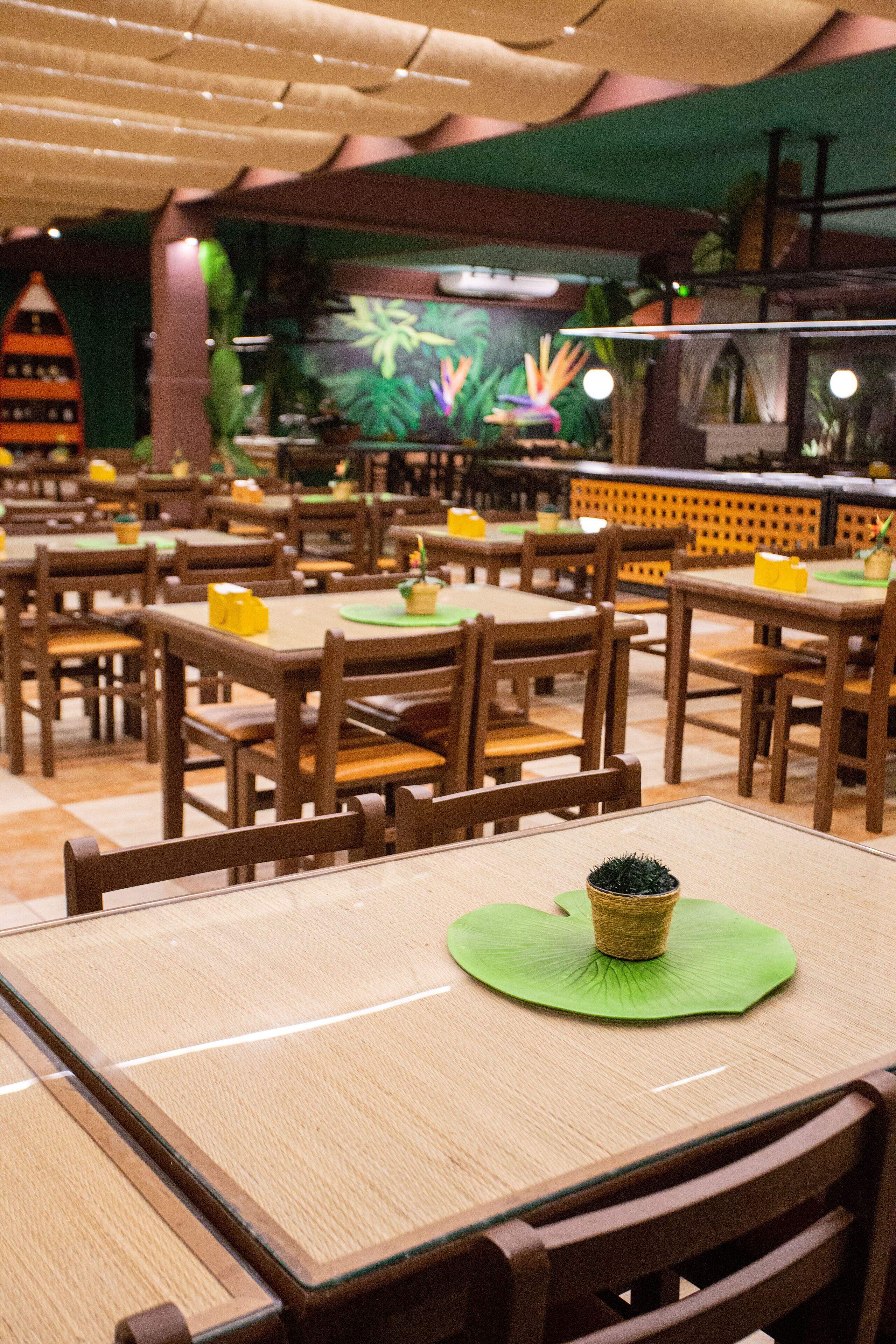 Interior de restaurante com mesas e cadeiras de madeira, jogos americanos verdes e um arranjo de planta decorativo.