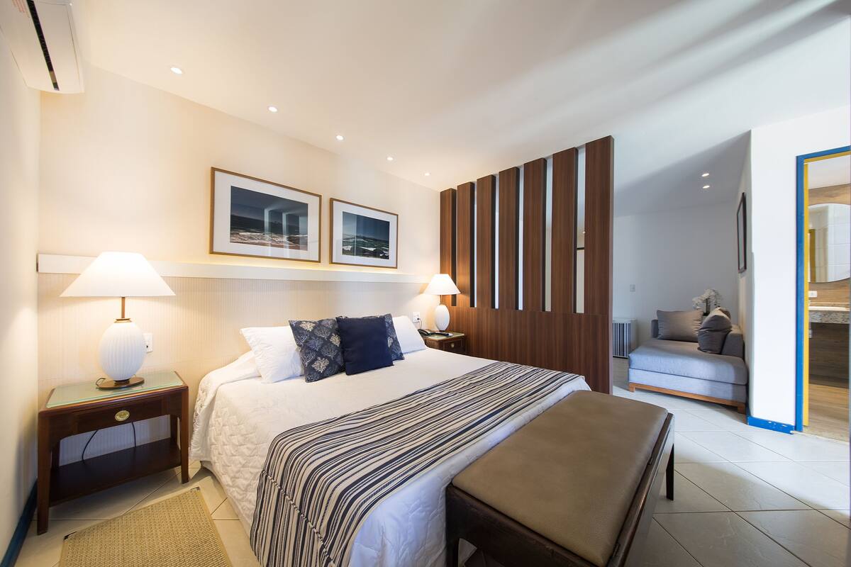A hotel room with a large bed, two bedside lamps, a wooden panel wall, a sofa, and two framed pictures on the wall.