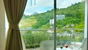 A balcony with a view of a green hillside and a building in the distance. There are two white plastic chairs and a small table with a vase of flowers. - P.S Hill Resort (Patong)