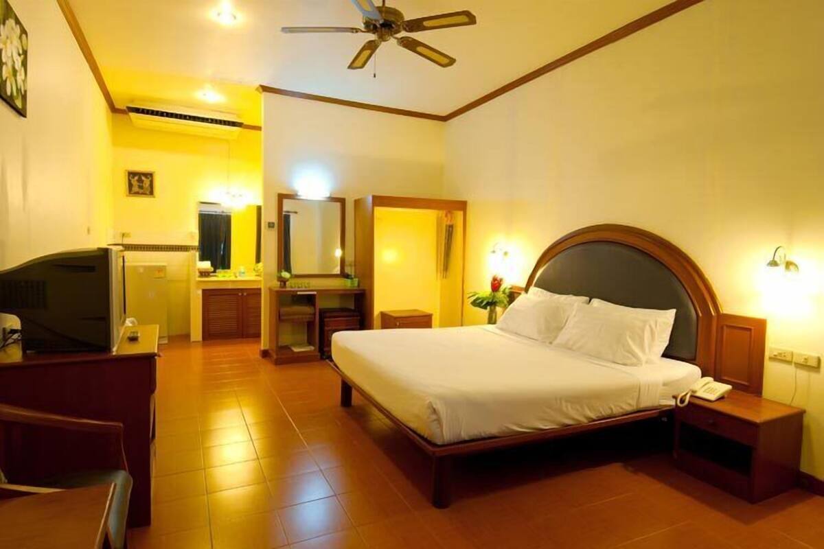 Habitación de hotel con cama, mesitas de noche, escritorio con silla, televisor, ventilador de techo y baño con espejo y lavamanos.