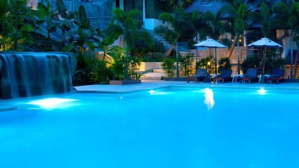 A pool with a waterfall feature, surrounded by lounge chairs and umbrellas, with a building in the background. - P.S Hill Resort (Patong)