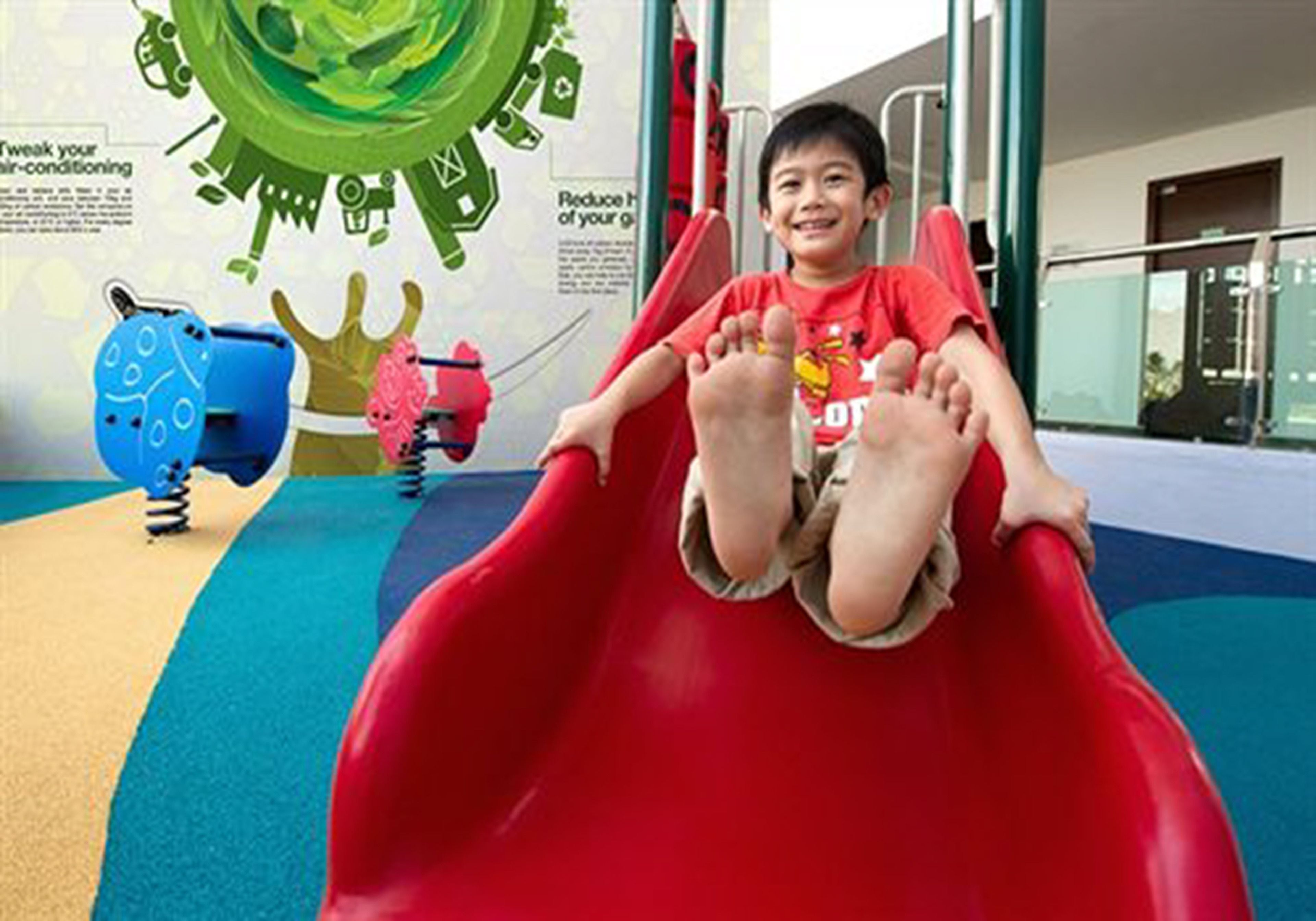 A child sliding down a red slide in a play area with colorful flooring and a green wall with educational graphics.