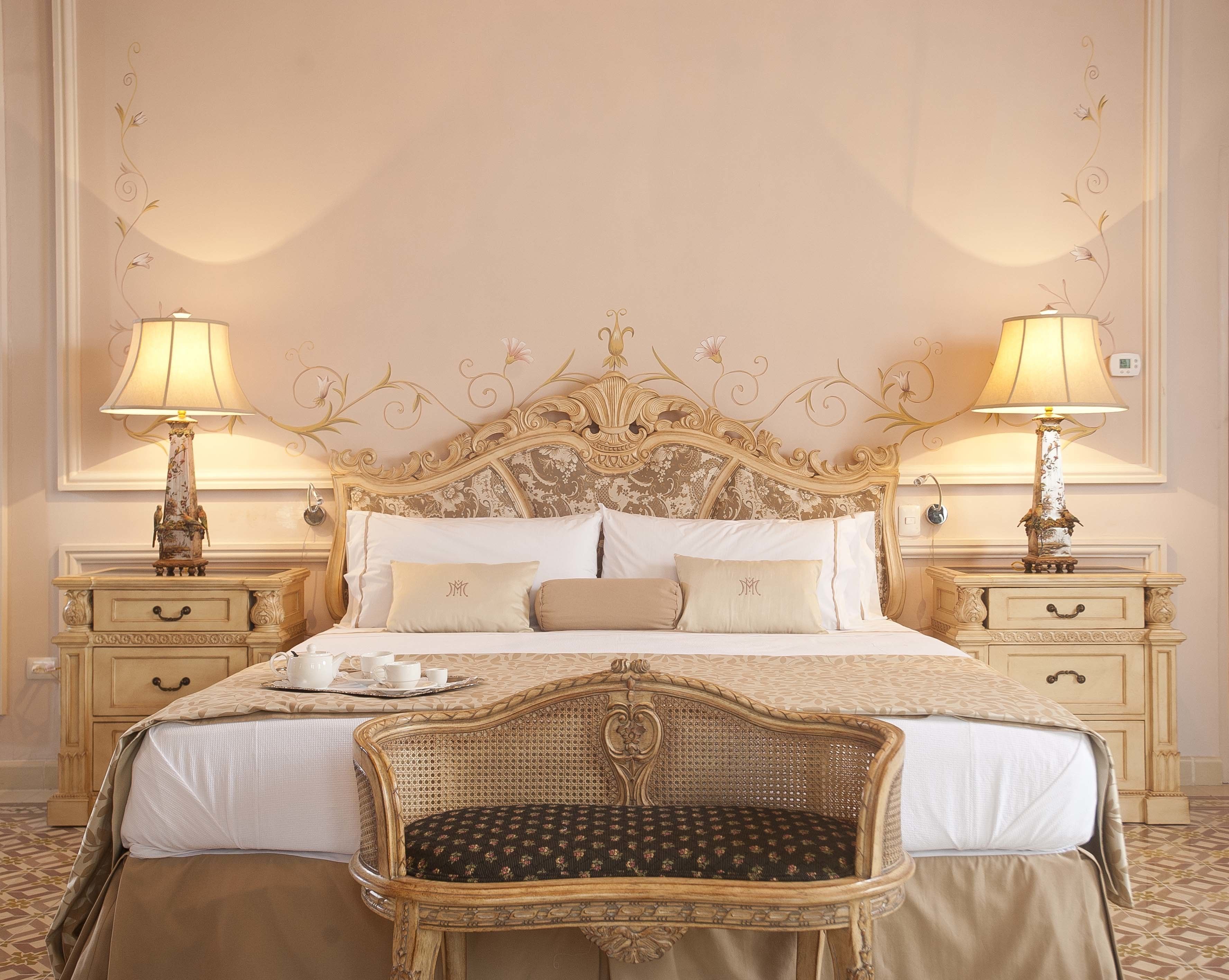 Camera d'albergo classica ed elegantemente arredata con un letto grande e decorato, due comodini con lampade, una poltrona in vimini e un vassoio con tazze e piattini.
