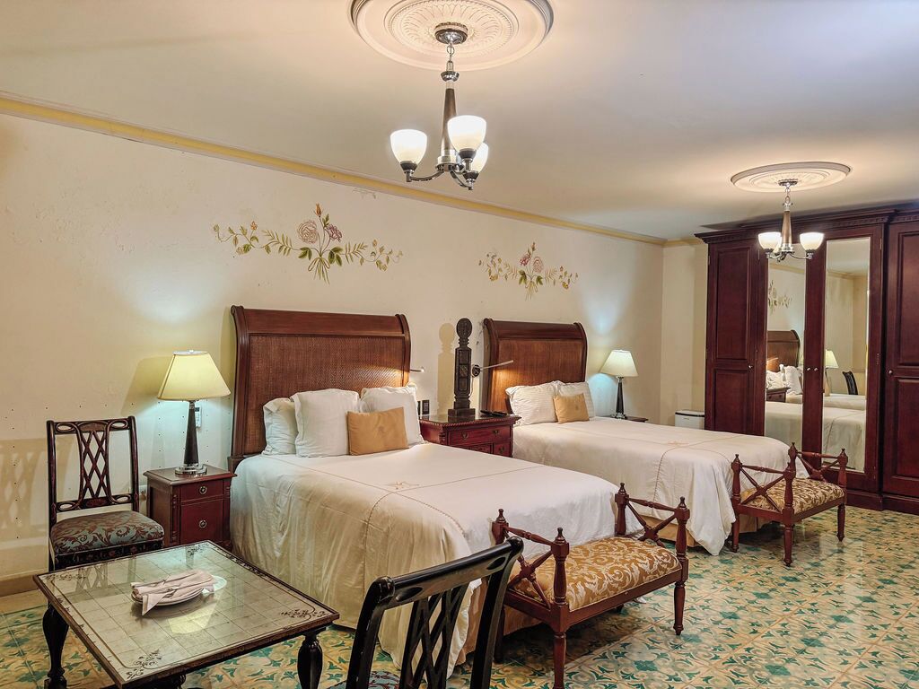 A hotel room with two beds, a table, chairs, a mirror, and a chandelier.