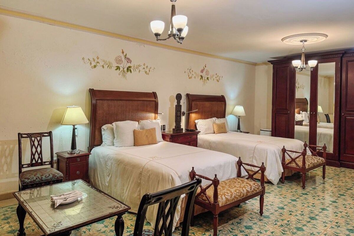 A hotel room with two beds, a table, chairs, a mirror, and a chandelier.