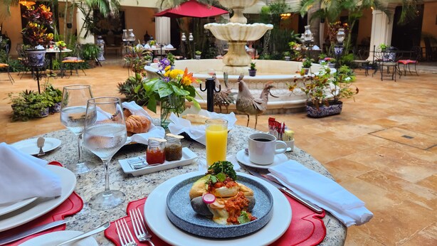 A dining table set with plates, glasses, and a variety of food items, surrounded by lush greenery and a fountain in a courtyard. - Mansión Merida Hotel Boutique - Restaurant  (Mérida)