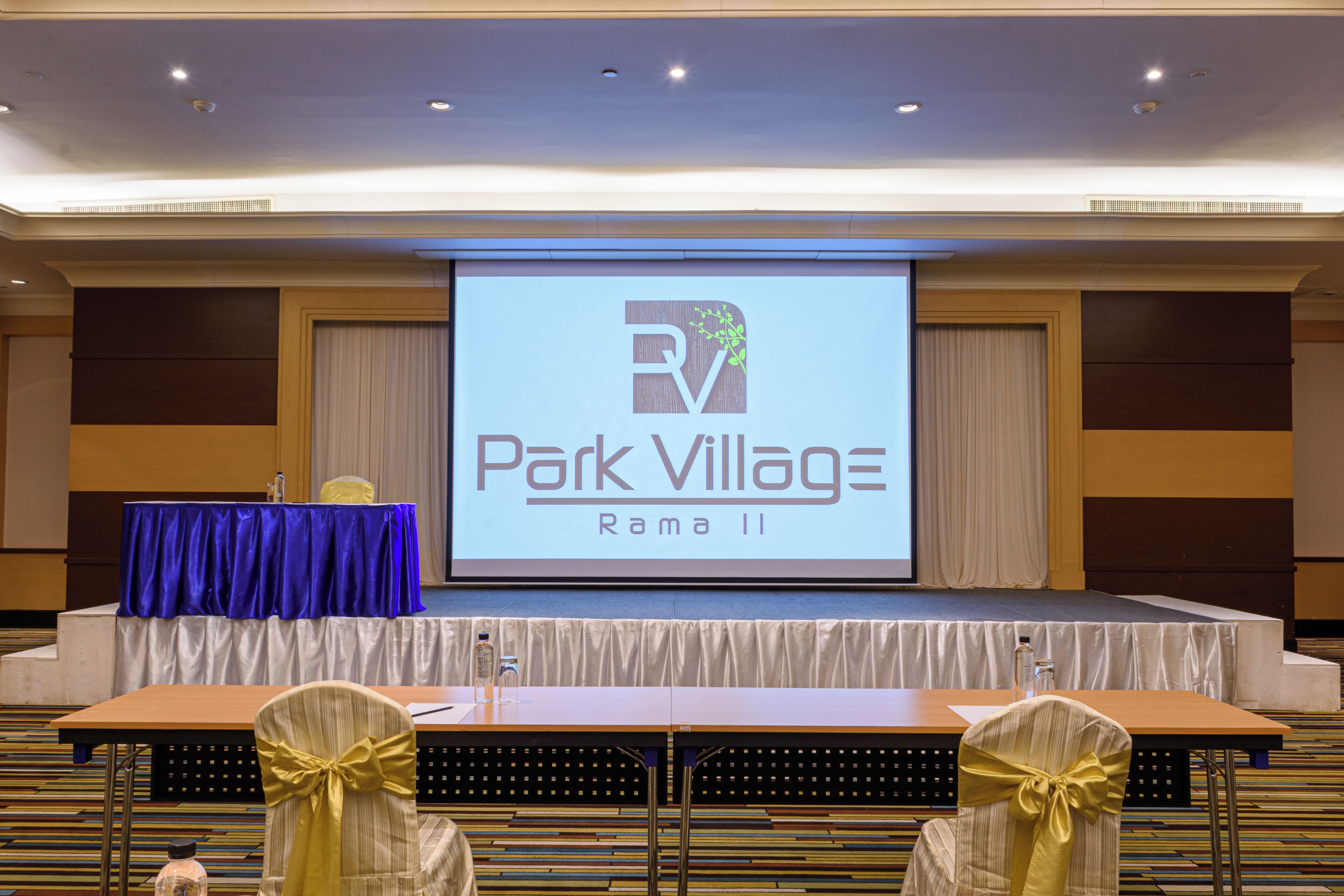 Ein Konferenzraum mit Bühne, Rednerpult und einer Leinwand, auf der 'Park Village Rama II' angezeigt wird.