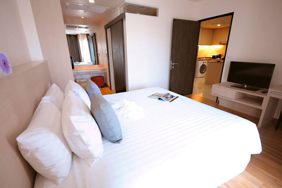 A hotel room with a bed, pillows, a TV, a desk, and a washing machine visible through an open door.