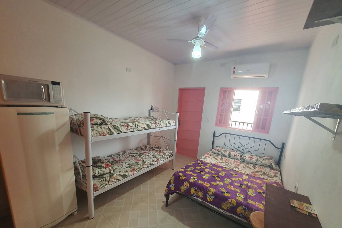Une chambre de dortoir équipée de lits superposés, d’un four à micro-ondes, d’un réfrigérateur, d’un ventilateur de plafond, d’une fenêtre avec un cadre rose et d’une petite table avec une chaise.