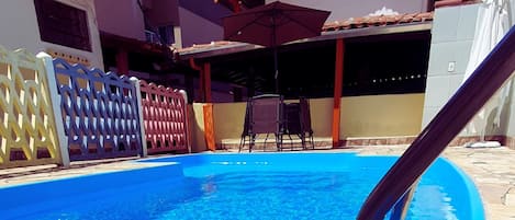 A swimming pool with a blue water surface, surrounded by a fence and a building with a tiled roof.