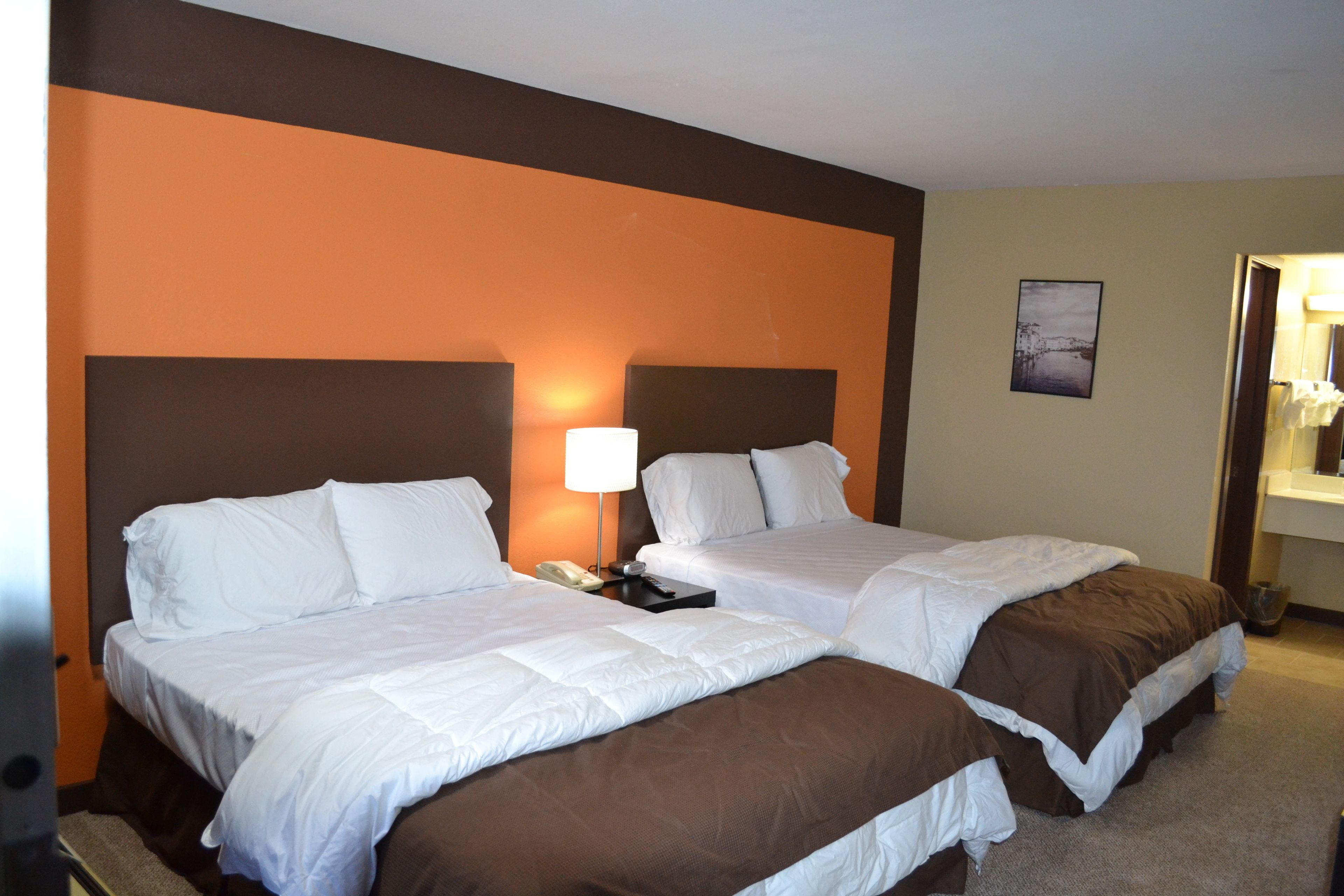 A hotel room with two beds, a nightstand with a lamp, a painting on the wall, and a bathroom visible through an open door.