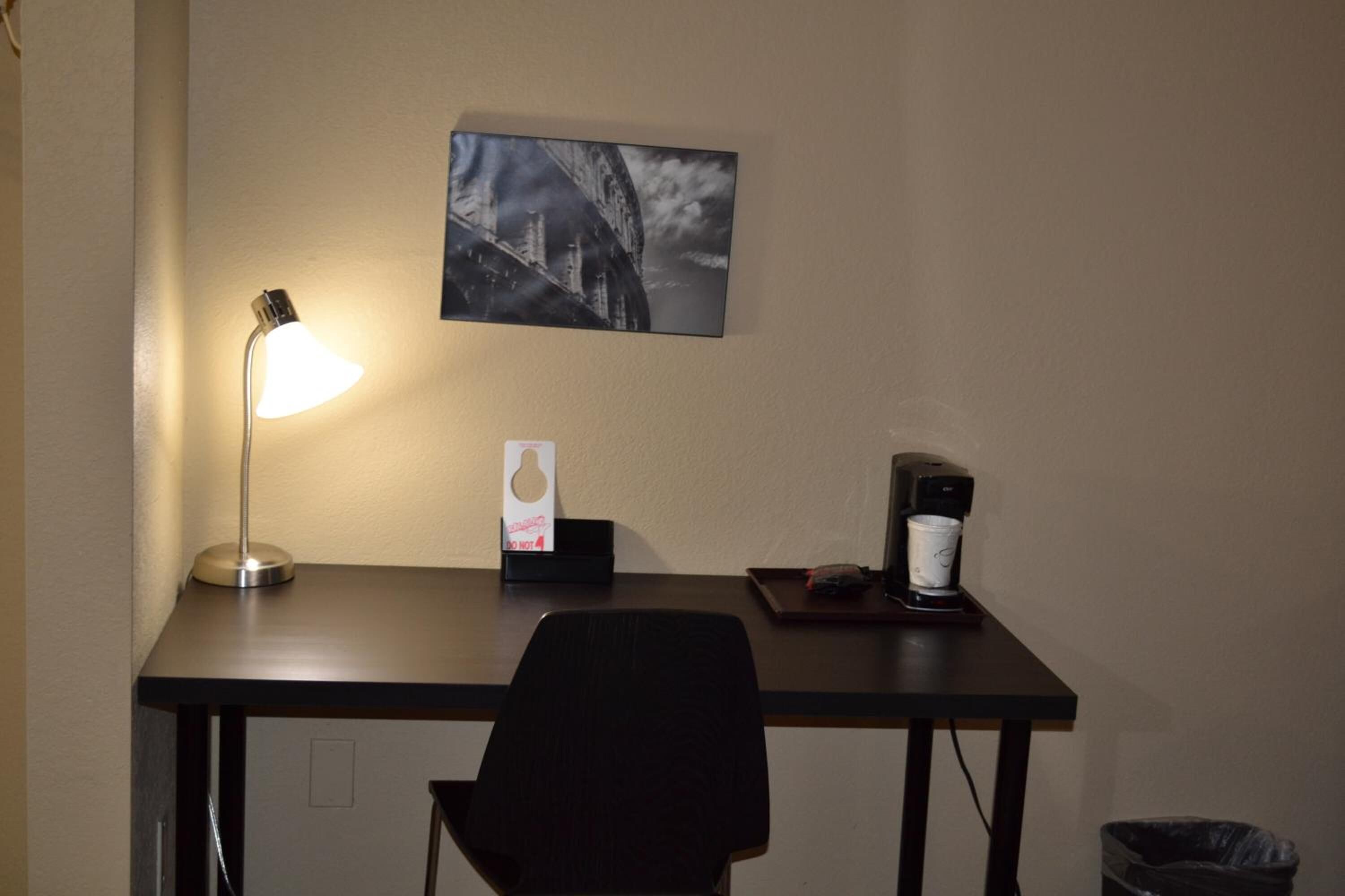 Ett hotellbord med en lampa, en kaffebryggare och en inramad tavla på väggen.