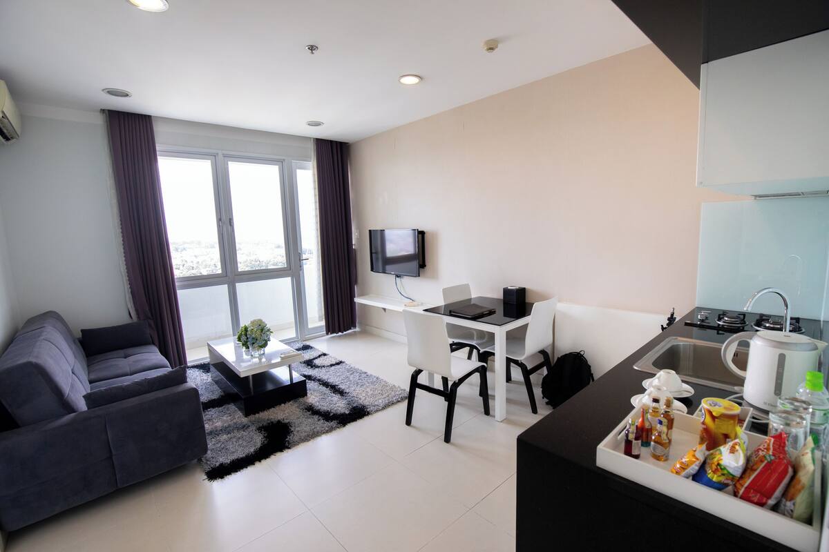 A modern hotel room with a sofa, a dining table, a TV, and a kitchenette.