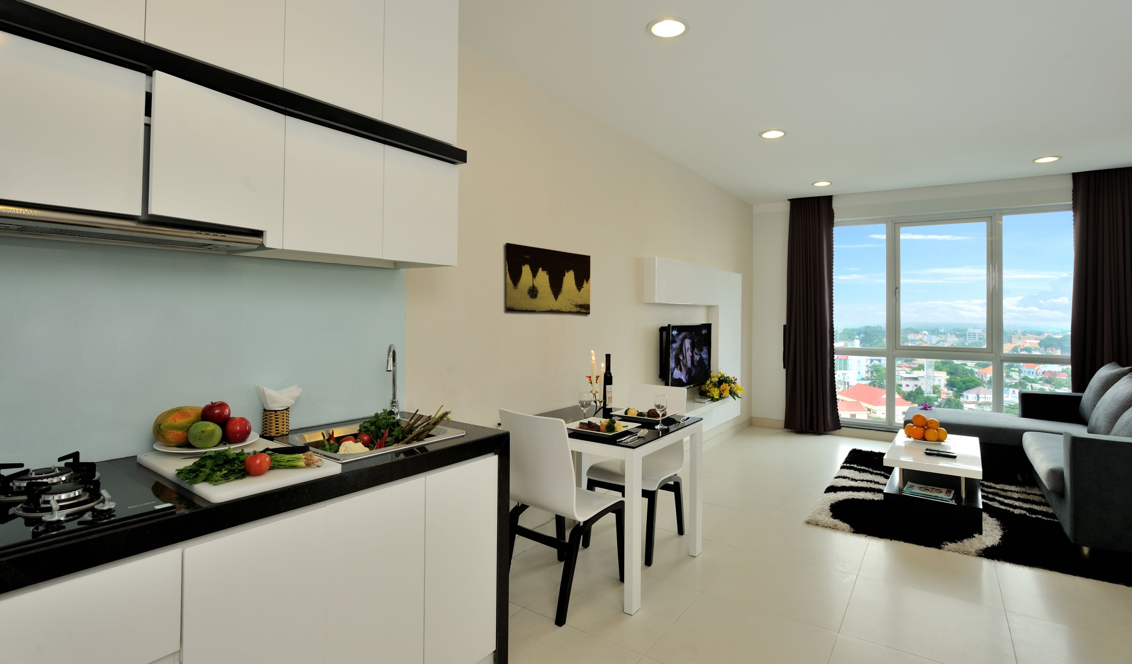 A modern apartment with a kitchen, dining area, and living room. It features a large window with a city view, a flat-screen TV, and a dining table with chairs.