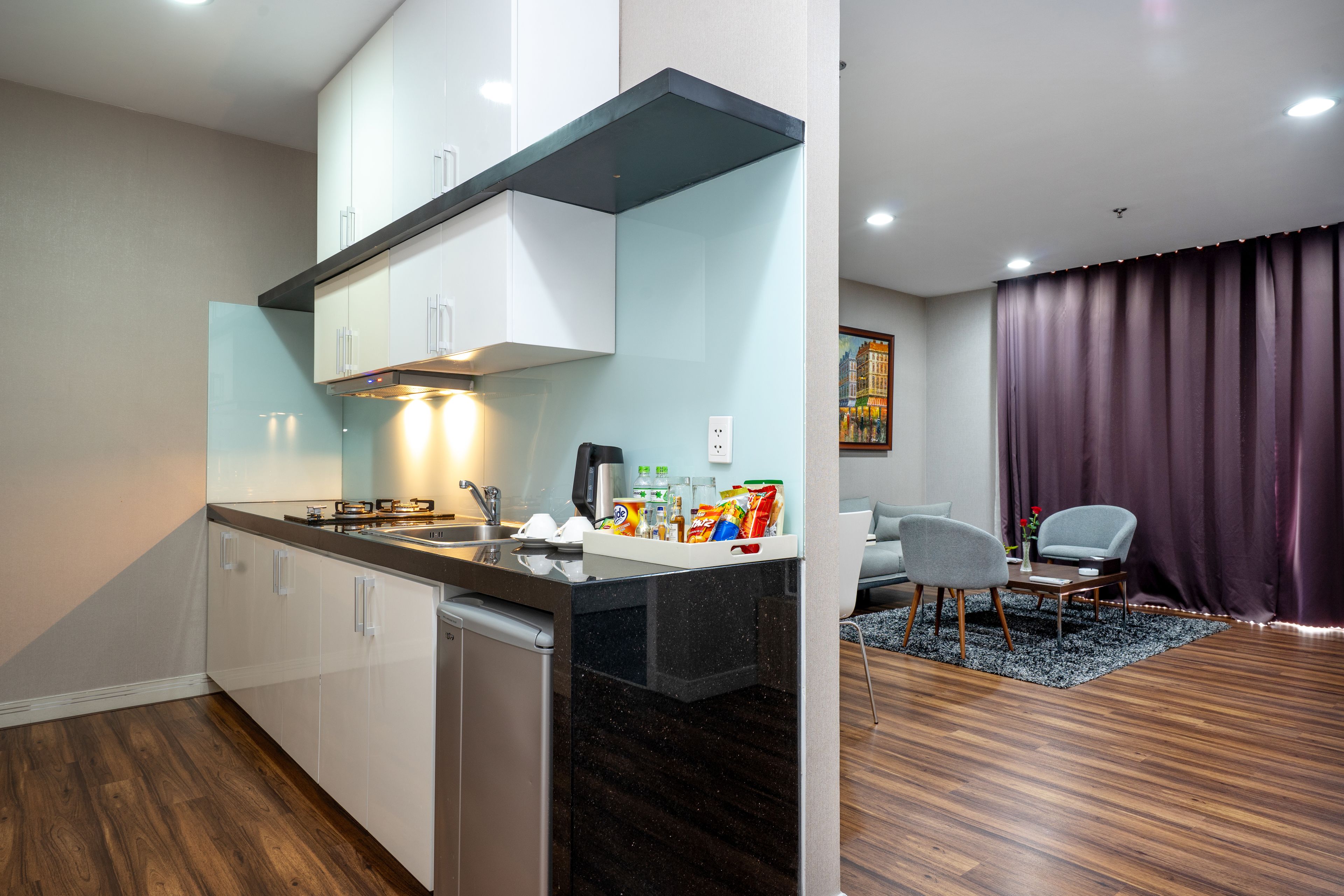 A modern kitchen with white cabinets, a black countertop, and a built-in dishwasher. There is a dining area with a gray chair and a table, and a purple curtain in the background.