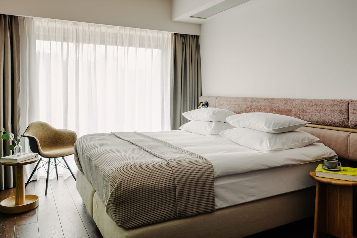 Ein modernes Hotelzimmer mit einem großen Bett, einem Holfnachtisch, einem Sessel und einem großen Fenster mit Vorhängen.