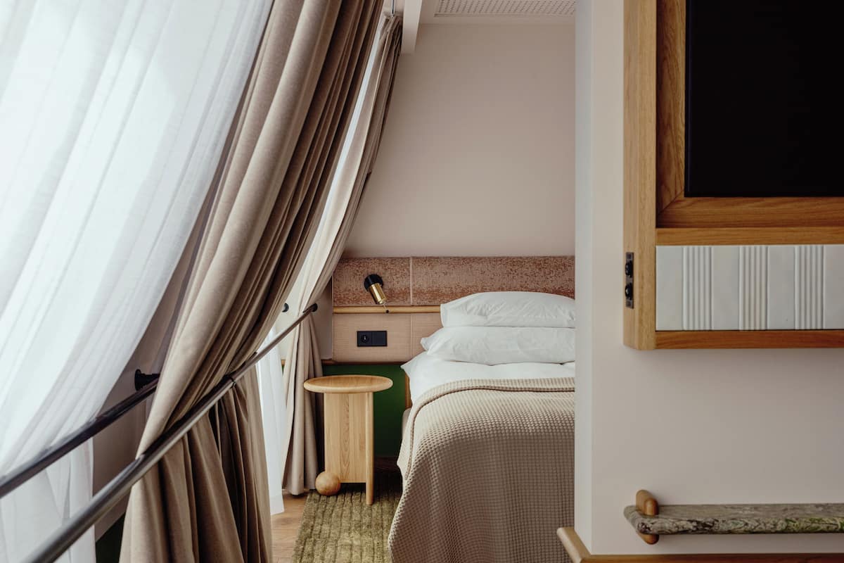 Ein Hotelzimmer mit Bett, Nachttisch, Lampe und Blick durch Vorhänge nach draußen.