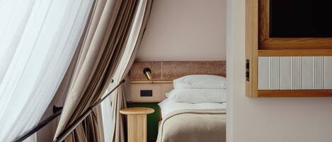 A hotel room with a bed, bedside table, lamp, and a view of the outside through curtains.
