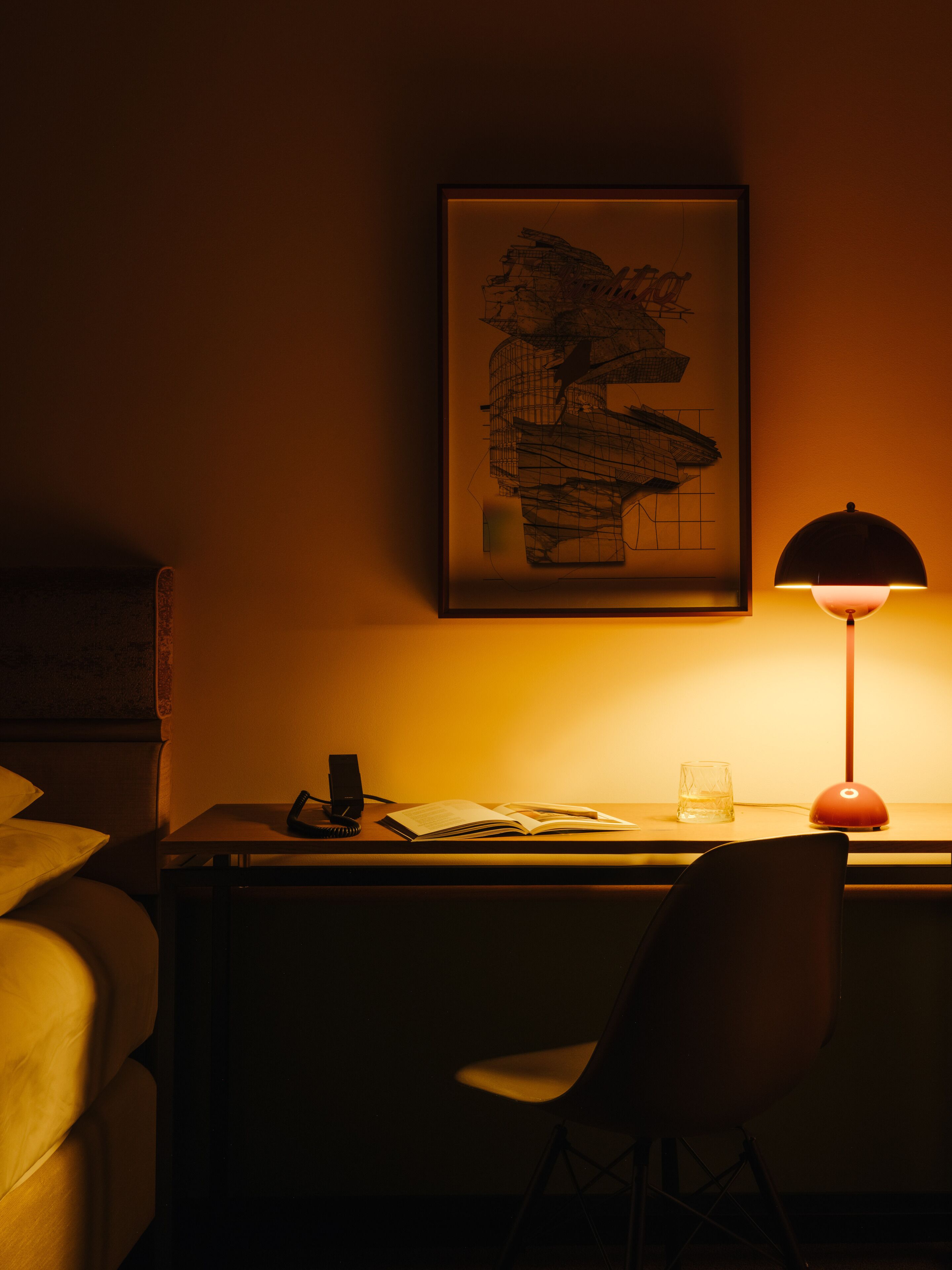 A dimly lit hotel room with a desk, a chair, a lamp, a framed artwork, and a bed.