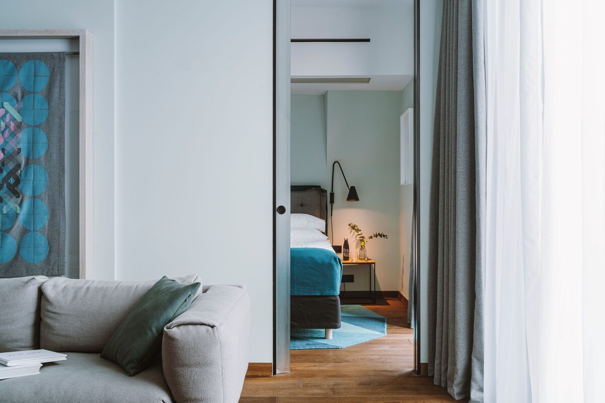 A modern bedroom with a bed, bedside table, lamp, and a view into another room.