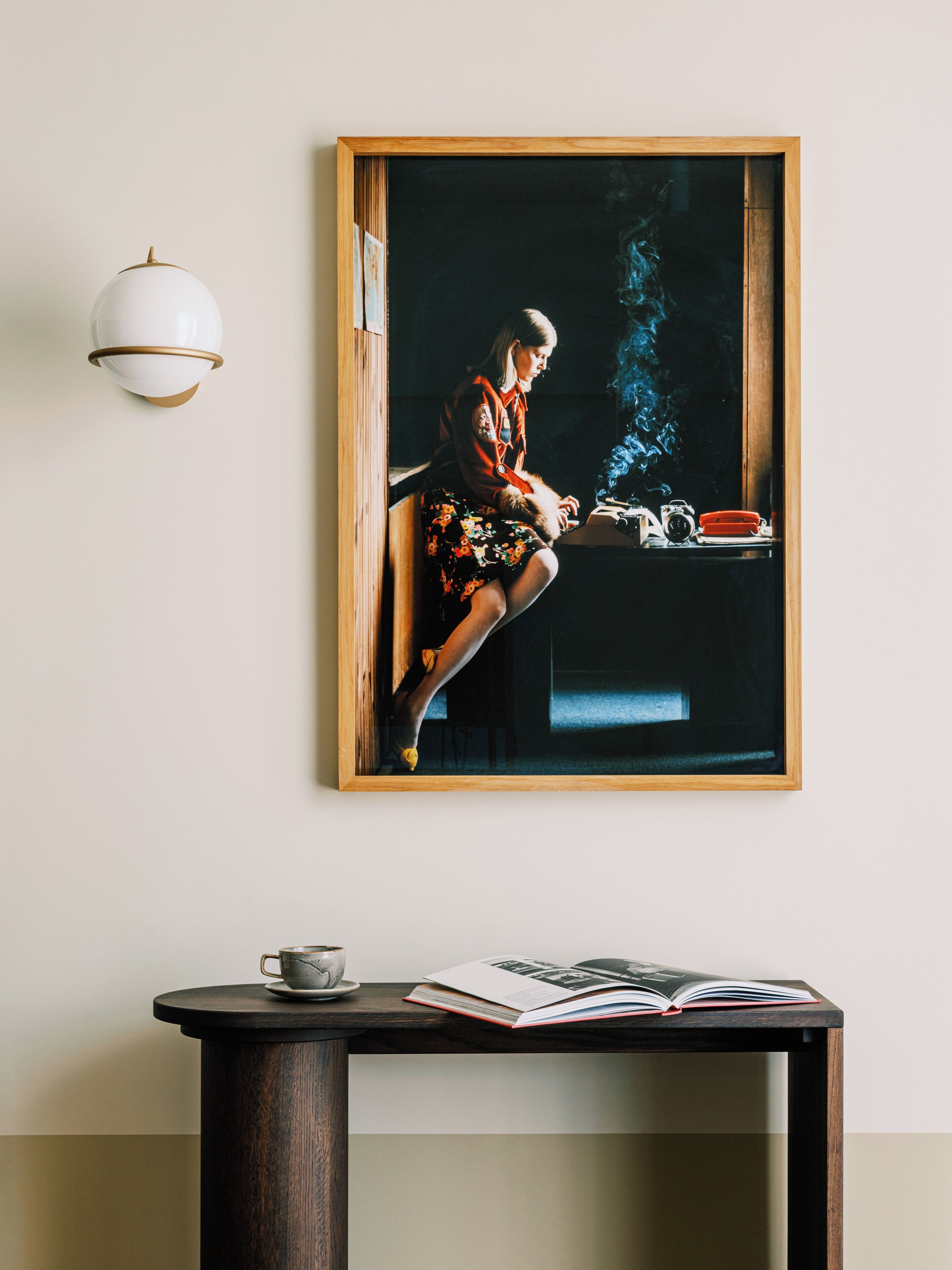 Ein gerahmtes Kunstwerk zeigt eine Person, die am Fenster sitzt, mit einem Rauch-Effekt, einen Tisch mit einem Buch und einer Tasse sowie eine Wandleuchte.