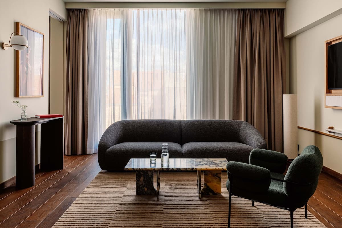 Ein modernes Wohnzimmer mit einem dunkelgrauen Sofa, einem Glastisch und einem grünen Sessel. Ein kleiner Beistelltisch trägt eine Vase und ein Buch. Große Fenster mit leichtem Vorhangstoff und schweren Vorhängen erhellen den Raum.