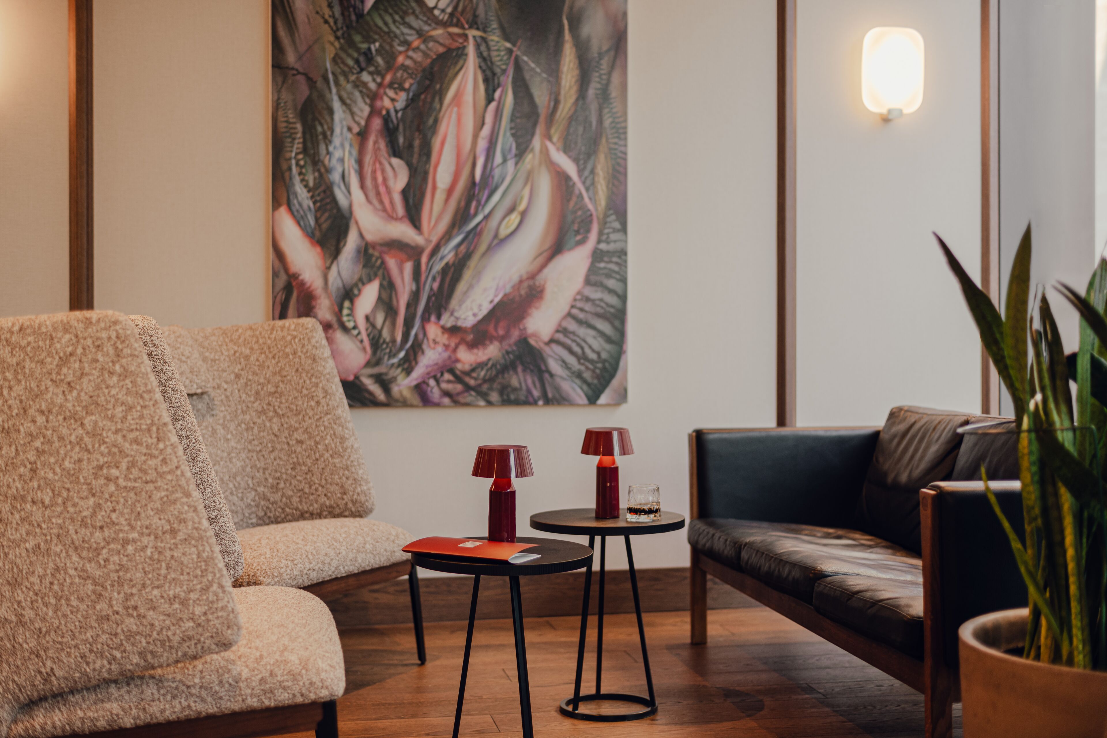 A modern living room with a beige armchair, a dark sofa, two small tables with red lamps, and a large floral painting on the wall.
