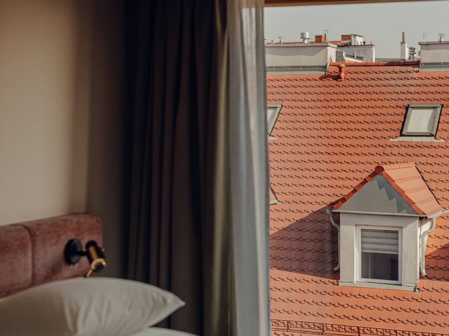 Habitación de hotel con cama, ventana con cortinas y vistas a edificios con tejados rojos.