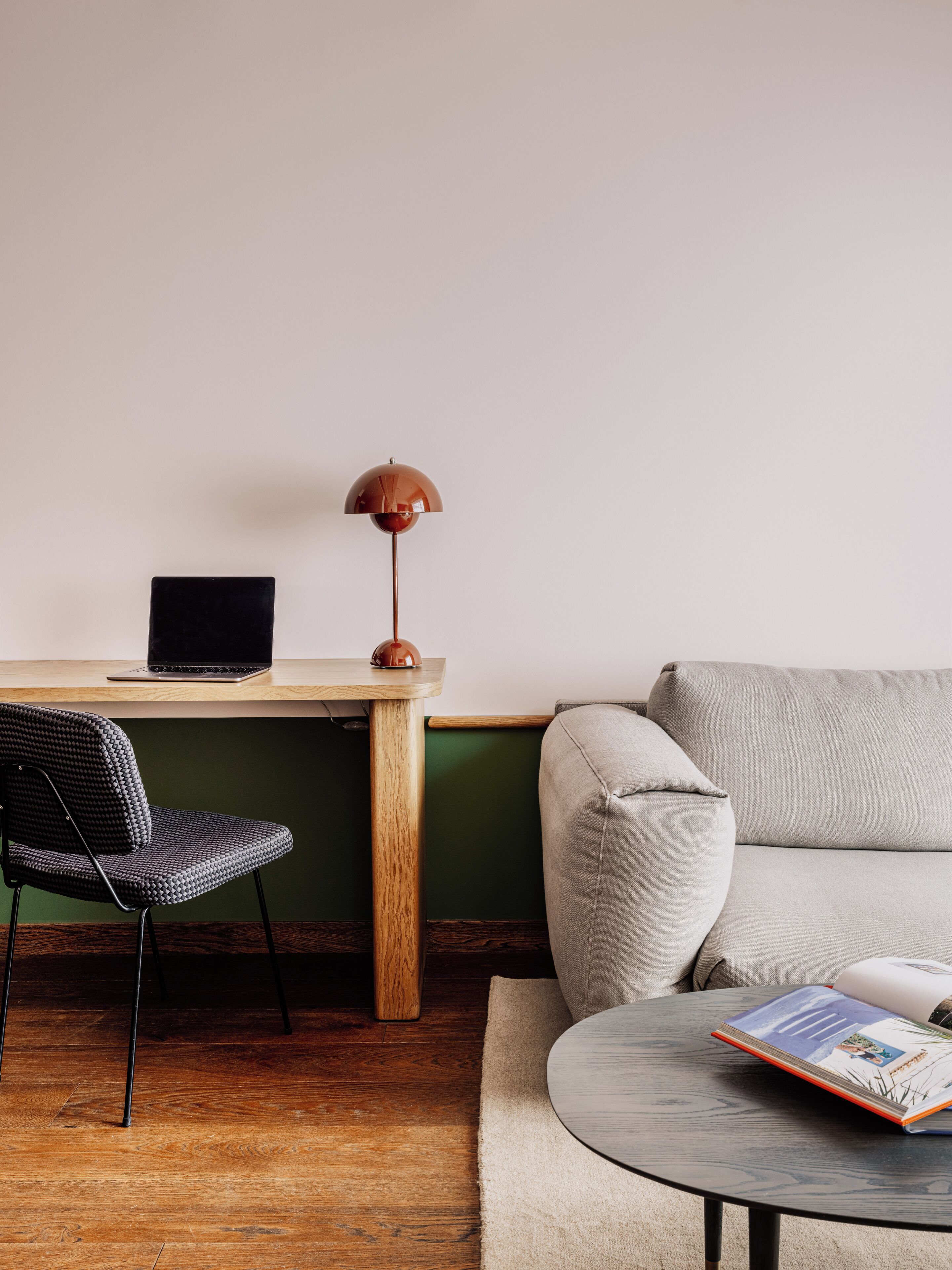 A workspace with a wooden desk, a laptop, a lamp, a chair, a sofa, and a small table with a book.