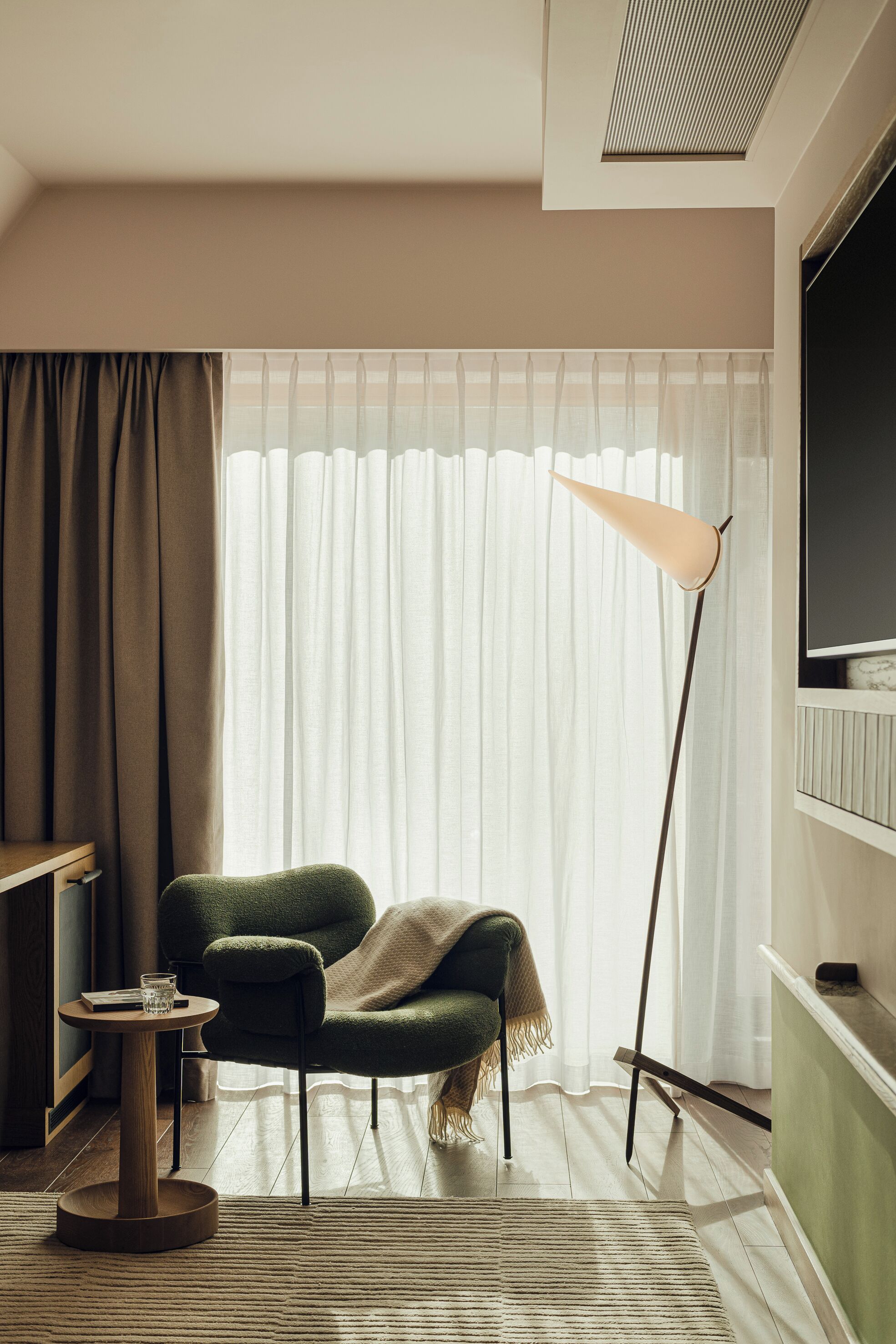 A room with a green armchair, a small round table with a glass, a floor lamp, and a large window with sheer curtains.