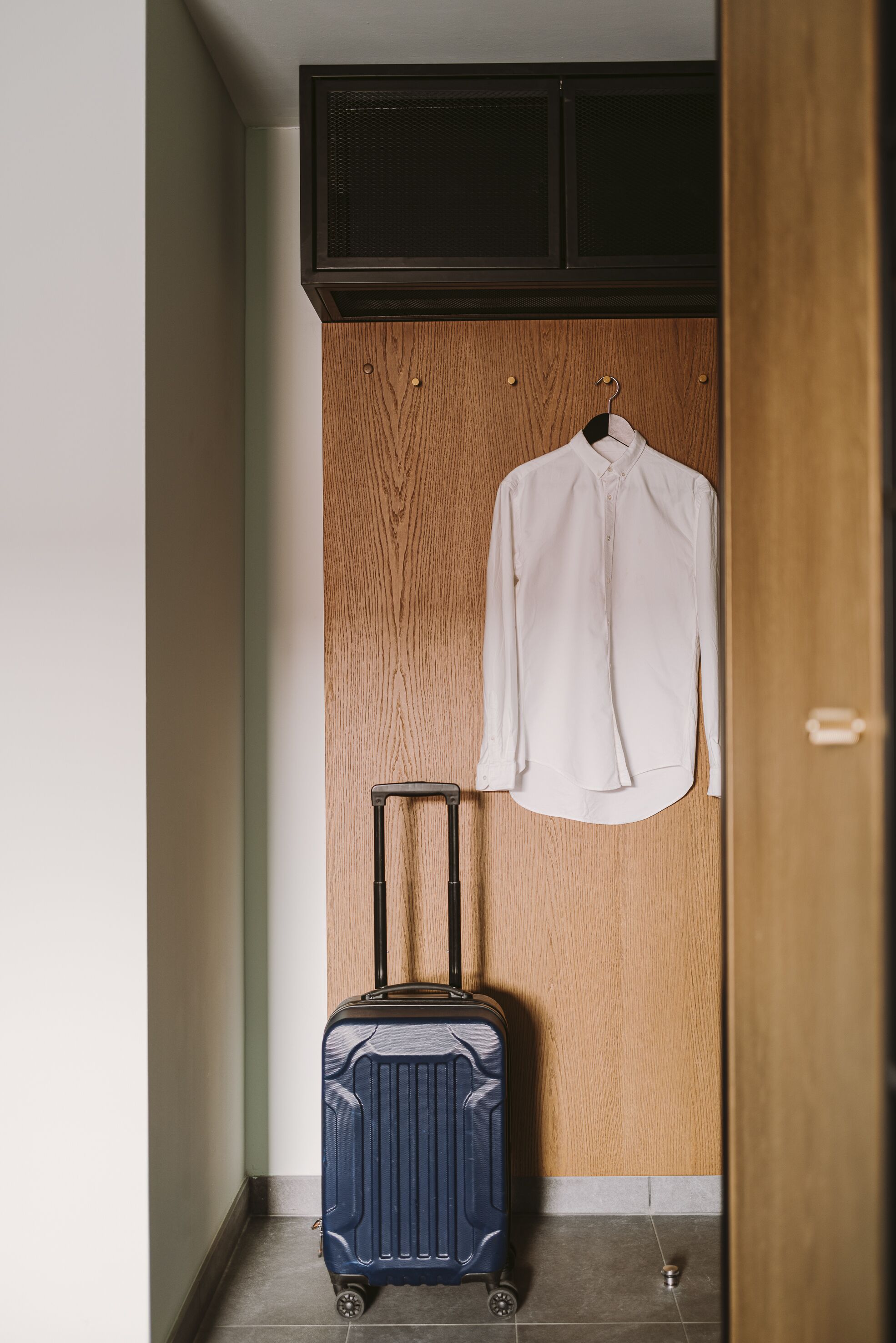 A room with a wooden wardrobe, a hanging white shirt, and a blue suitcase on the floor.