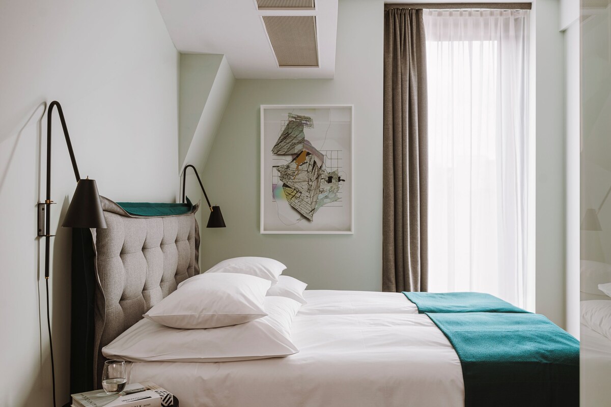 Un lit bien fait, avec du linge de lit blanc et un plaid turquoise, une lampe de chevet et une œuvre d’art abstraite encadrée accrochée au mur.