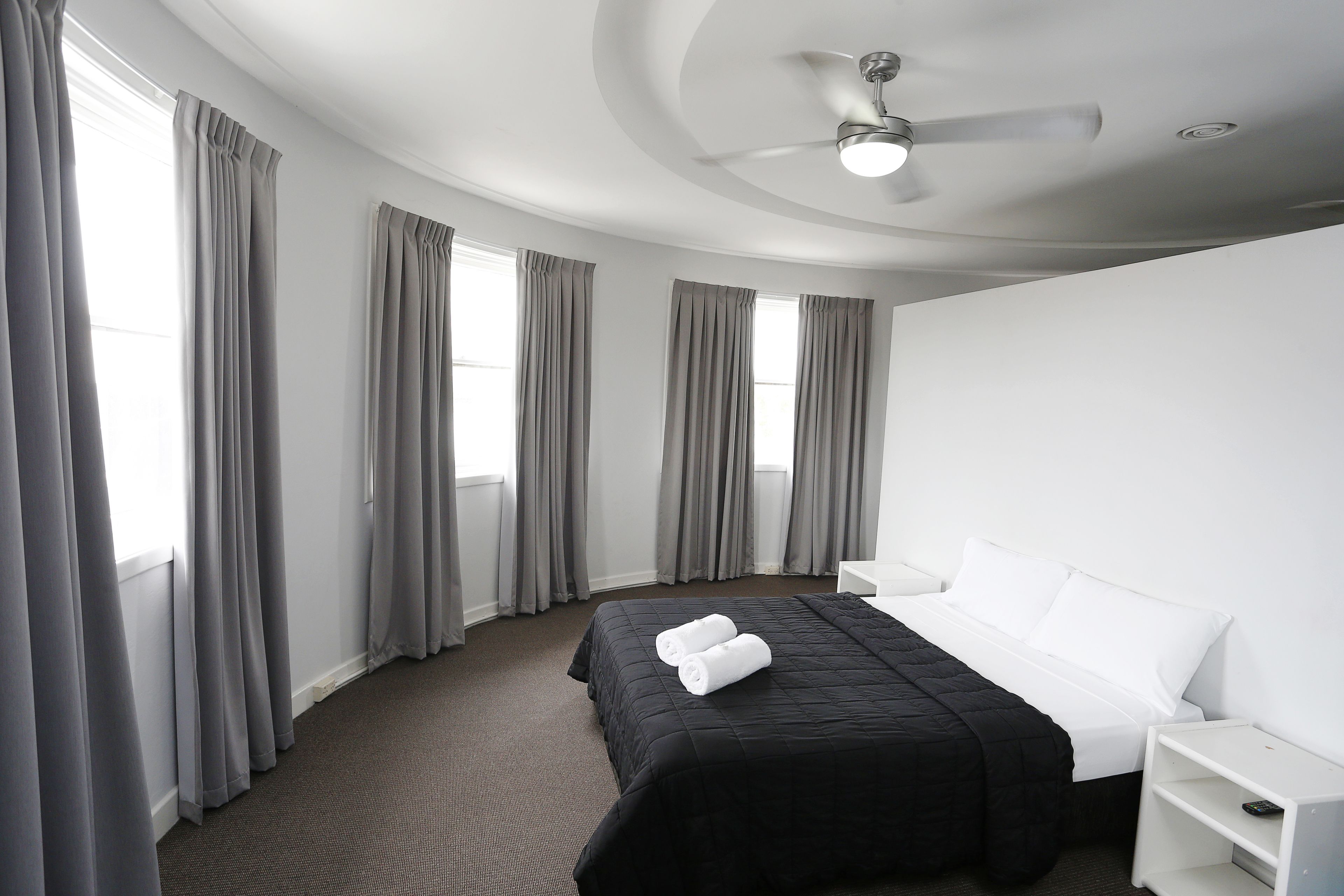 Habitación de hotel con cama, mesita de noche blanca y ventilador de techo.