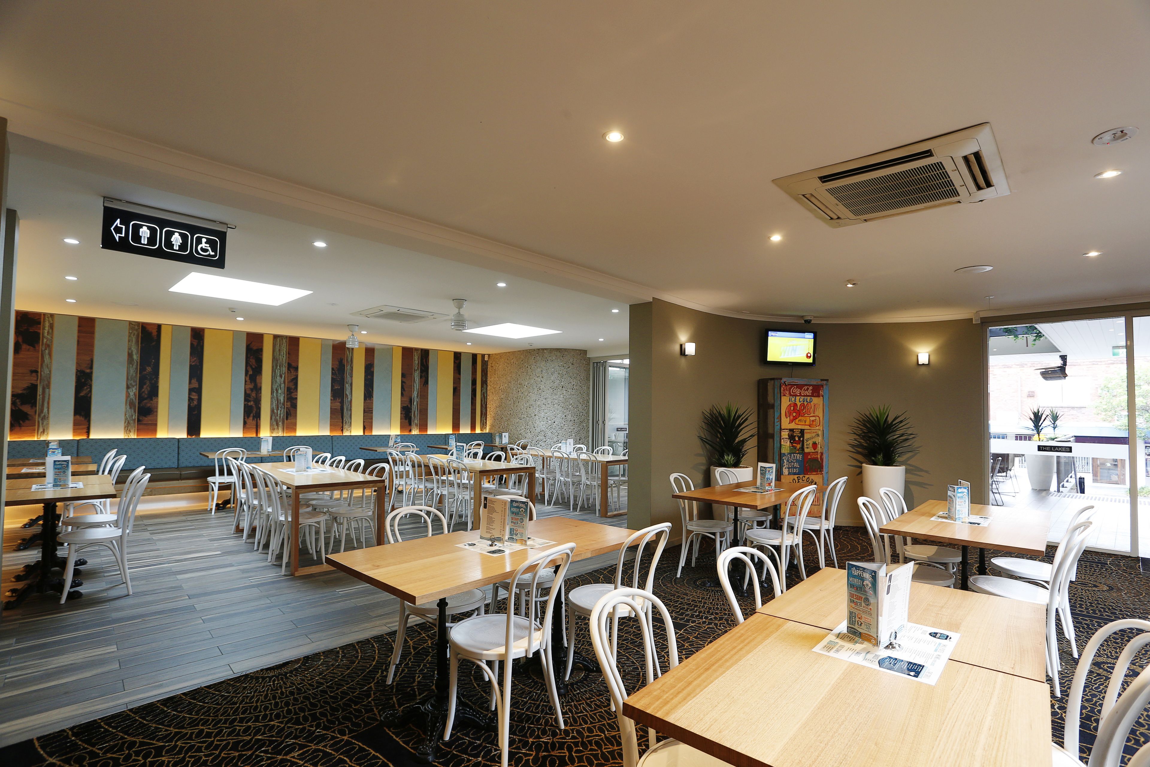 A hotel dining area with multiple tables and chairs, a sign indicating amenities, and a television screen.