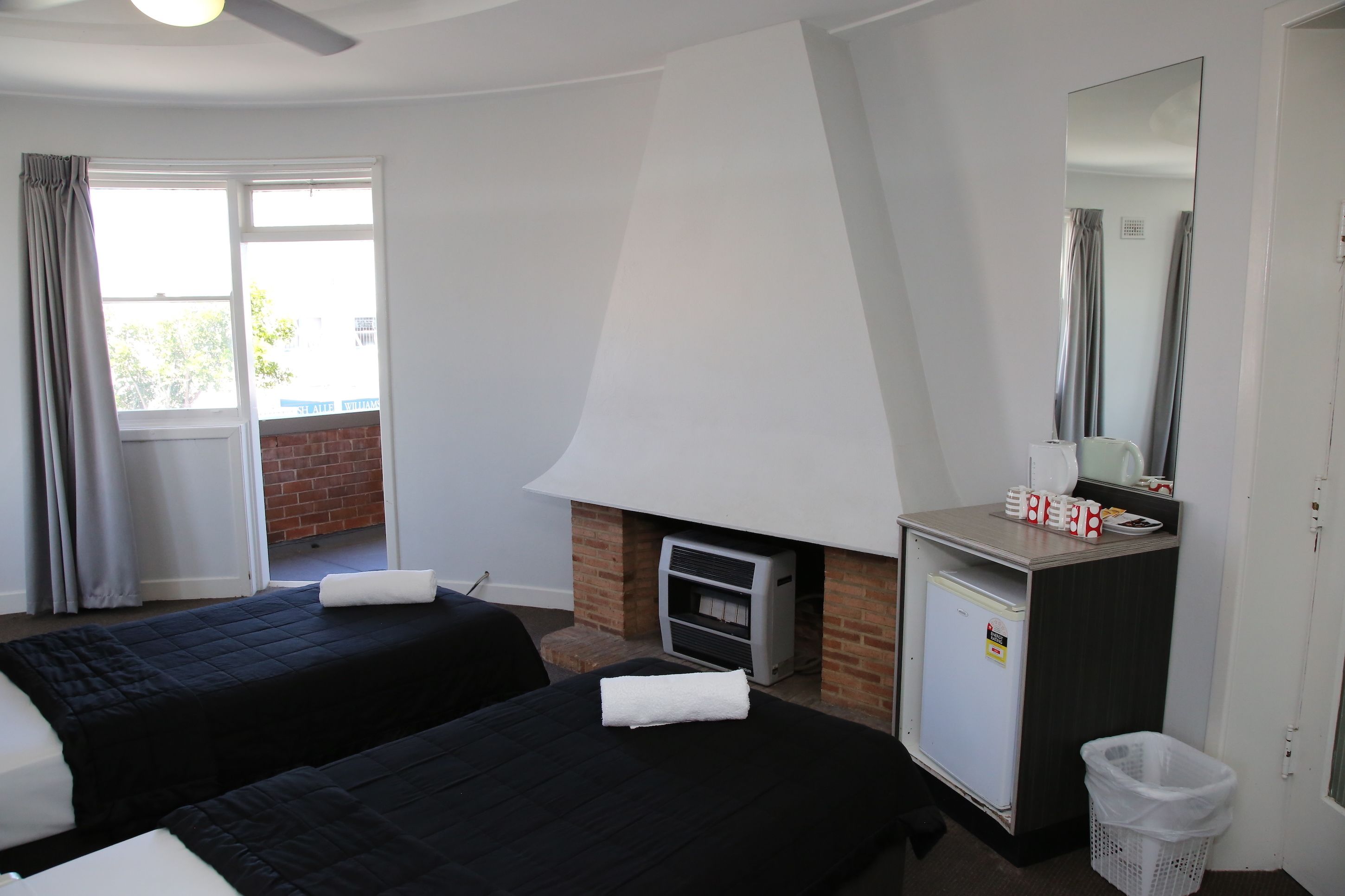 A hotel room with two beds, a small kitchenette, a mirror, and a window with curtains.