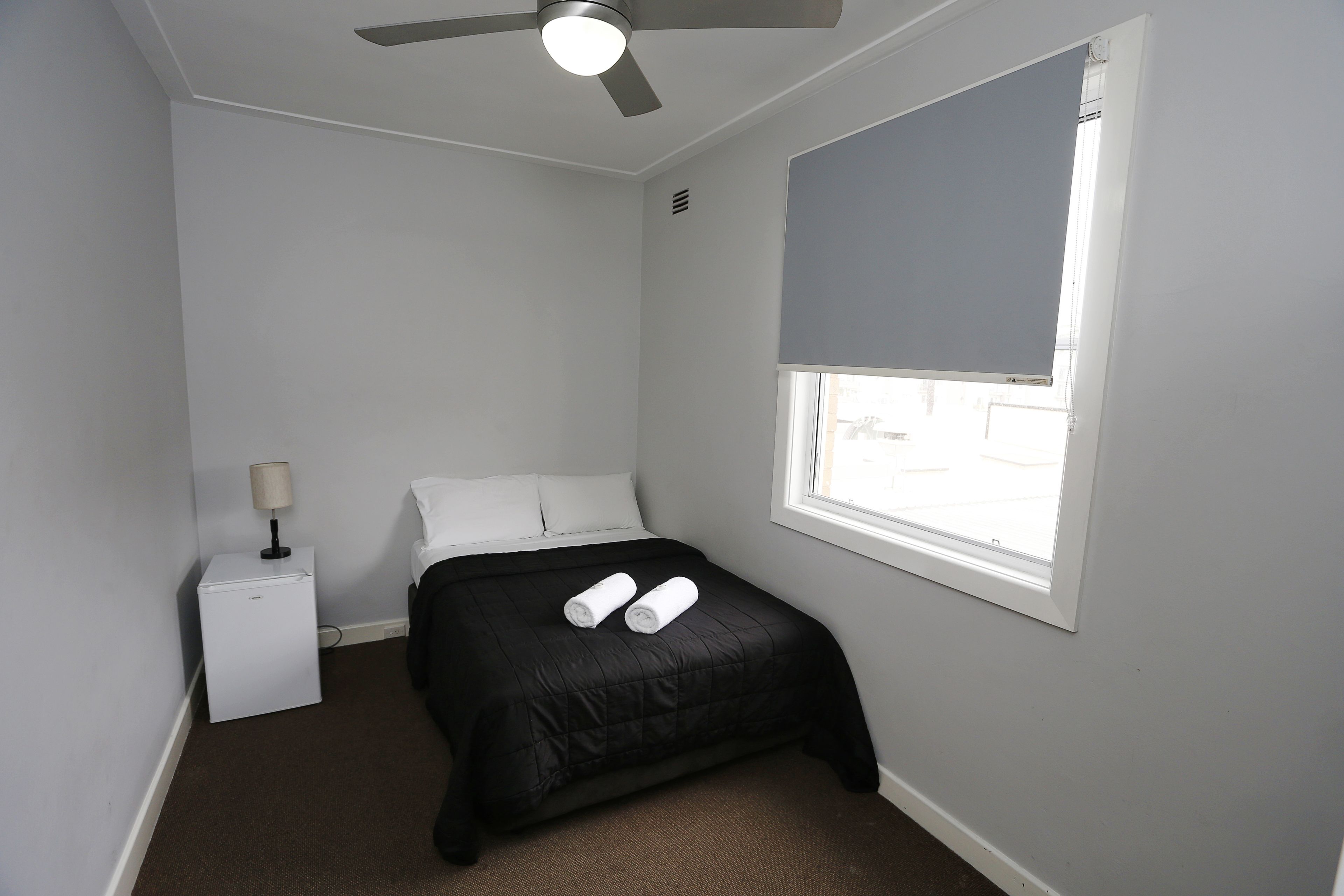 A small, neatly arranged bedroom with a single bed, a nightstand, a lamp, and a ceiling fan.