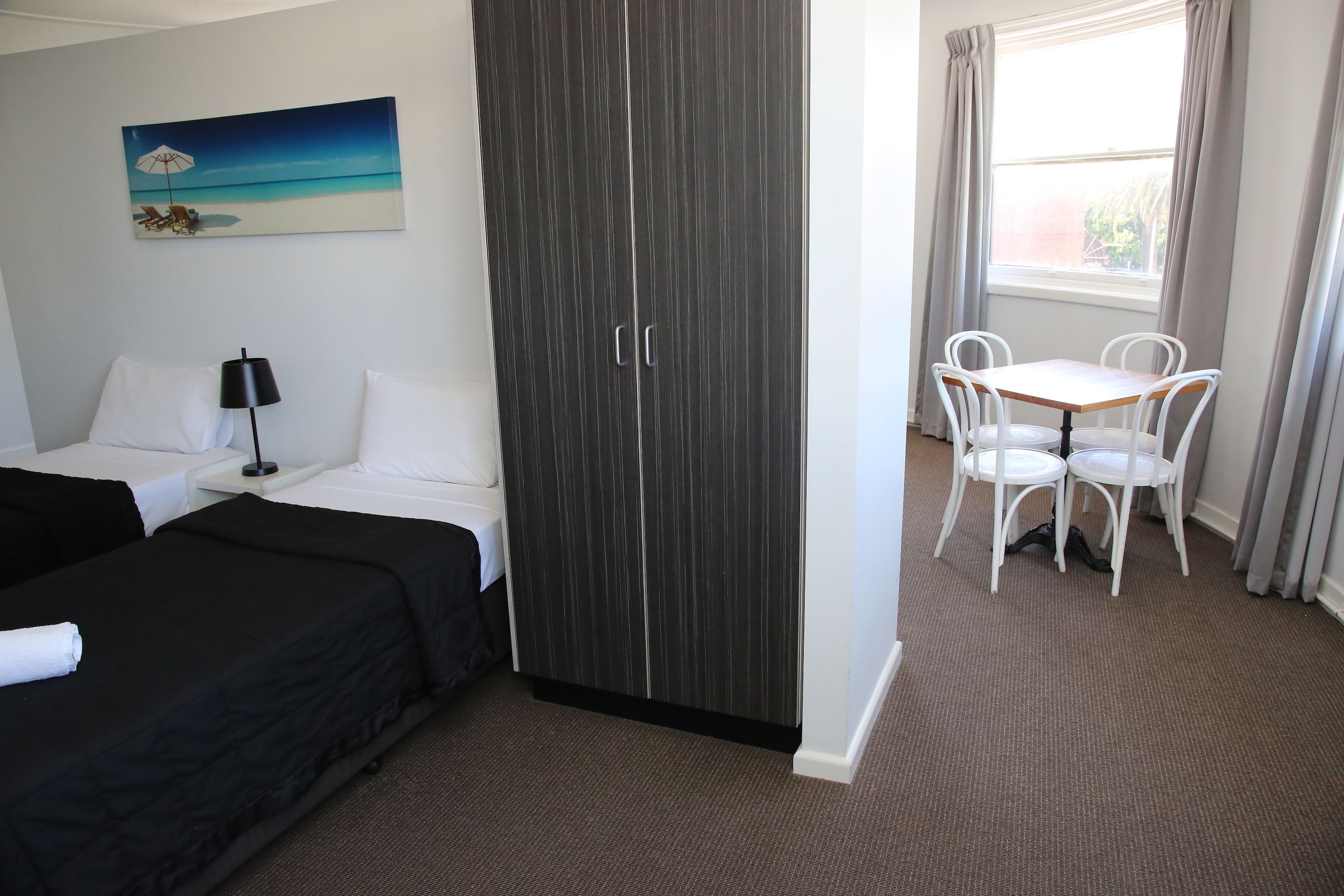 A hotel room with two beds, a wardrobe, a small table with chairs, and a window with curtains.