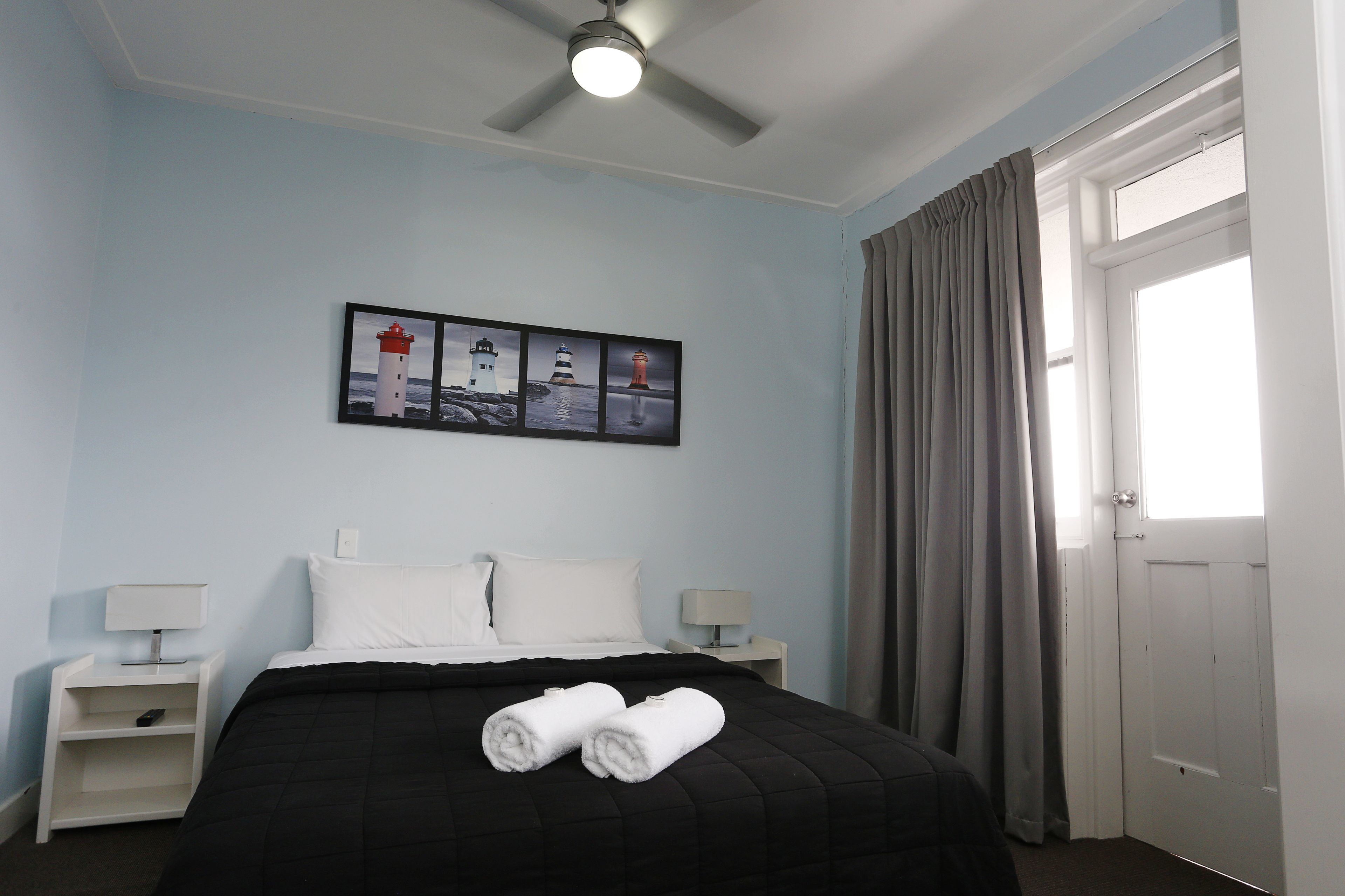 Ein Hotelzimmer mit einem Bett, zwei Nachttischen mit Lampen, einem Deckenventilator, einem Fenster mit Vorhängen und einem an der Wand hängenden Bild von Leuchttürmen.