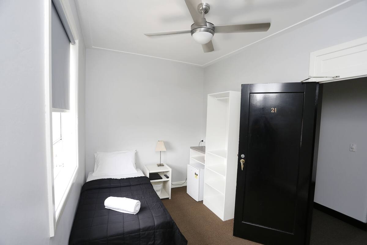 A small, clean room with a bed, a desk, a wardrobe, and a ceiling fan.
