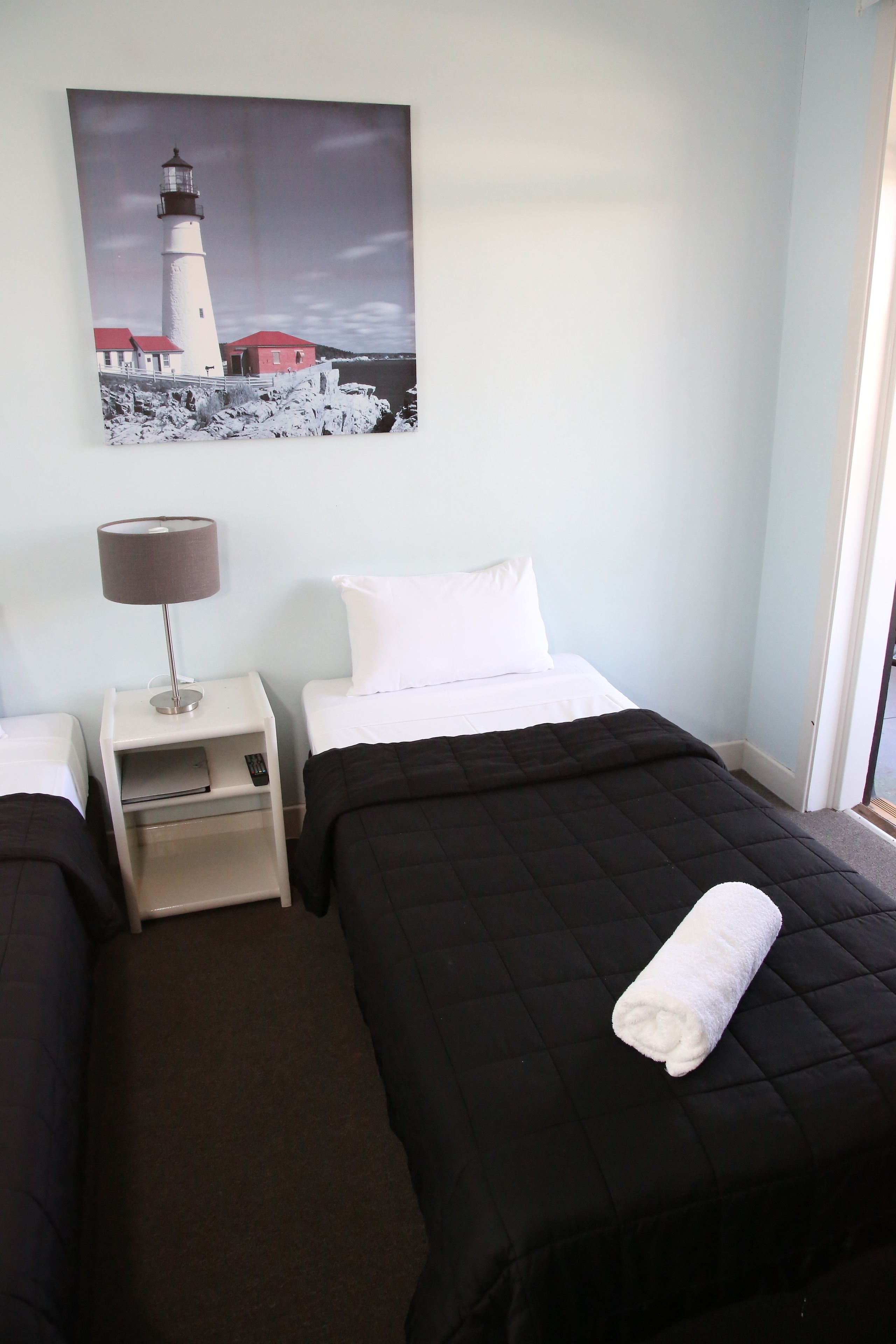 A hotel room with two beds, a nightstand with a lamp, a painting of a lighthouse, and a view of the sea.