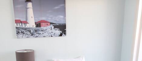 Habitación de hotel con dos camas, una mesita de noche con lámpara, un cuadro de un faro y vista al mar.