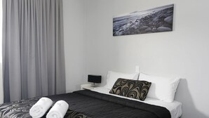 A neatly made bed with a patterned bedspread, two rolled towels, a black bedside lamp, a pillow with a swirl pattern, and a painting of a rocky beach on the wall. - The Lakes Hotel (The Entrance)