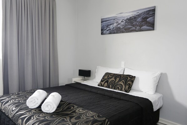 A neatly made bed with a patterned bedspread, two rolled towels, a black bedside lamp, a pillow with a swirl pattern, and a painting of a rocky beach on the wall.