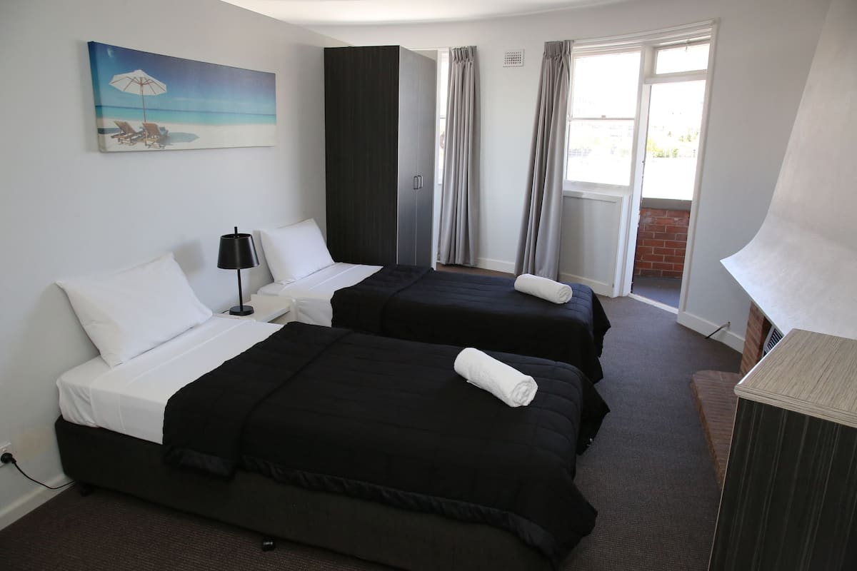 A hotel room with two beds, a painting of a beach scene, a desk with a lamp, and a view of the outdoors through large windows.